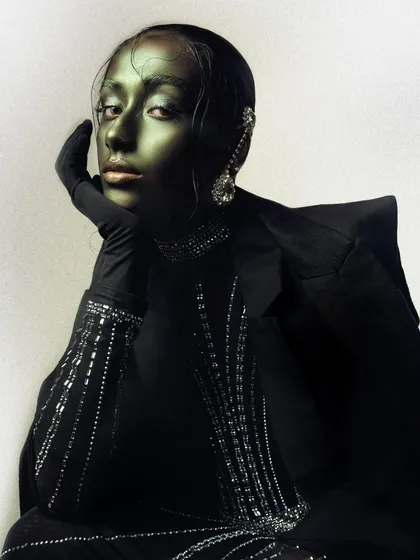 From a beauty story titled 'Chrome,' this image uses metallic green body paint and slicked-back hair to create a non-human, statuesque effect. The lighting is carefully controlled to highlight the sheen and dimension of the makeup.