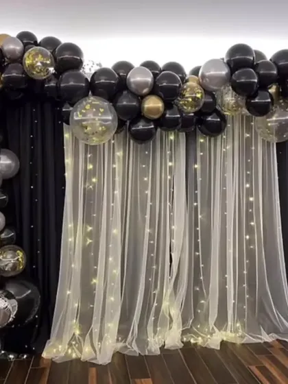 A classic black and gold balloon arch against a curtain backdrop with fairy lights, suitable for any elegant party or reception.