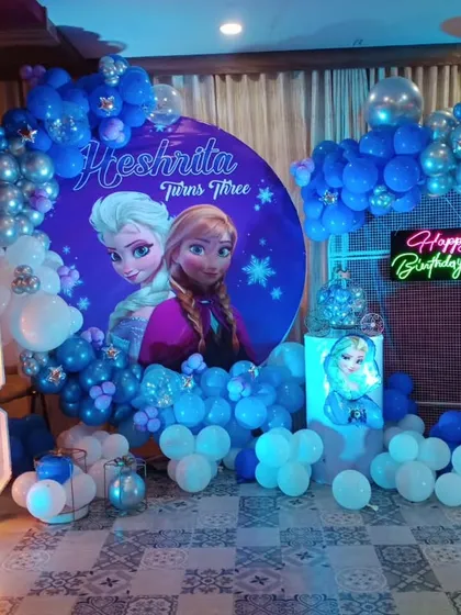 A magical Frozen themed birthday party, with a blue and silver balloon arch, character backdrops of Elsa and Anna, and a light-up marquee number '3'.