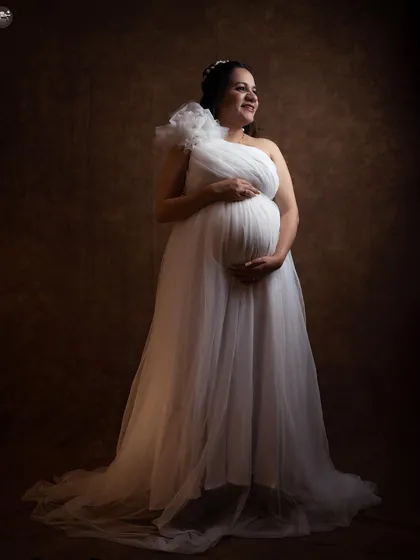 A classic and elegant solo portrait from a studio session. The soft lighting and the beautiful white one-shoulder gown create a timeless, angelic look that focuses on the joy of the mother-to-be.