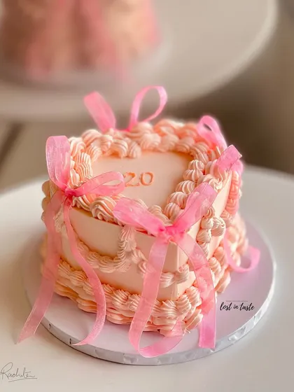 A beautiful heart-shaped cake in a soft pink hue, decorated with classic vintage Lambeth piping and delicate organza ribbons. This design is perfect for celebrating a 20th birthday in style.