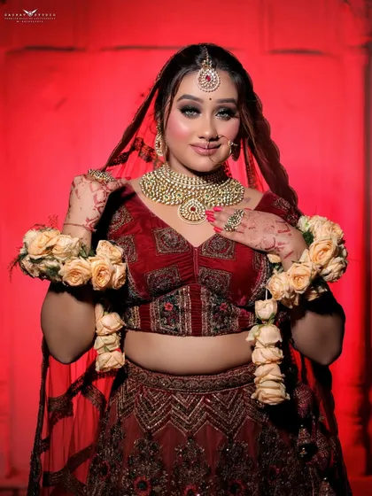 A bride in a deep red velvet lehenga, holding floral garlands (varmala), ready for a wedding-themed shoot.