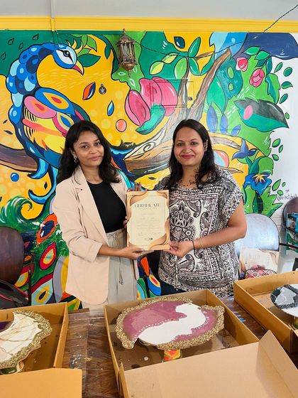 An instructor presents a certificate to a participant who created a lovely purple and gold resin art piece, with our vibrant peacock mural in the background.