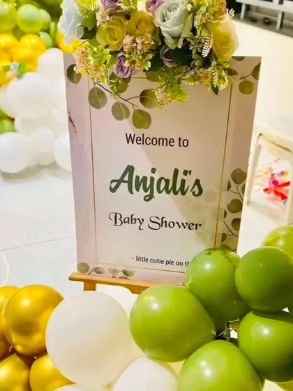 A close-up of a personalized welcome sign for 'Anjali's Baby Shower'. The elegant sign is placed on an easel and surrounded by a lush arrangement of green, white, and gold balloons.