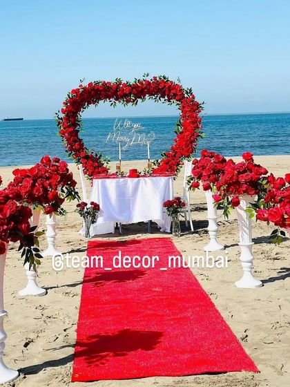 The stunning beach proposal scene, with the red rose arch perfectly framing the "Will you Marry Me?" sign against the blue sky and sea.
