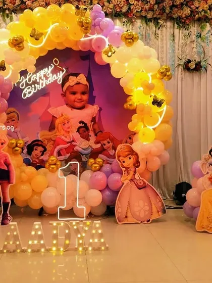 A fairytale first birthday celebration with a Disney Princess theme. This setup includes a circular backdrop with a photo, a warm-toned balloon arch, and cutouts of Cinderella, Snow White, and other beloved princesses.