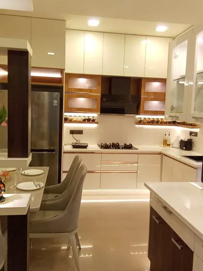This L-shaped modular kitchen is designed for both beauty and function. The combination of off-white and wood-finish cabinets, frosted glass shutters, and under-cabinet lighting creates a warm, inviting space for cooking and gathering, complete with a cozy breakfast counter.