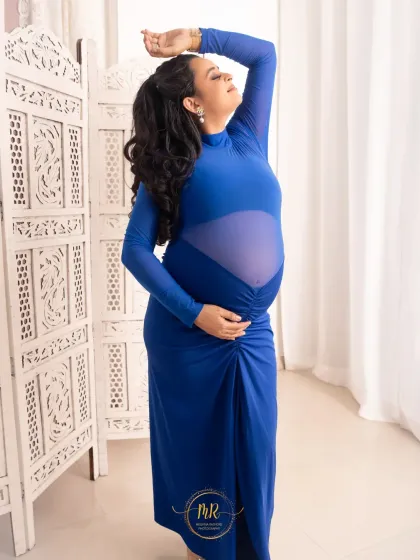 A full-length portrait showcasing a stylish, modern blue maternity dress. The pose is confident and elegant, set against a bright, airy studio backdrop with a decorative screen.