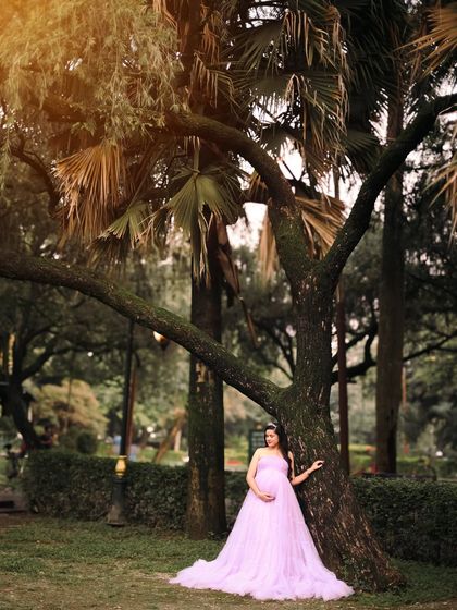 A wide shot of the mother-to-be in a beautiful lavender gown, standing gracefully by a tree. This image captures the grandeur of nature and the beauty of pregnancy.