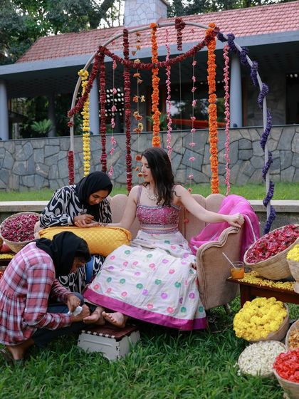 The bride gets her henna done, seated on a beautiful couch against a backdrop of hanging marigold garlands for her outdoor Mehendi.