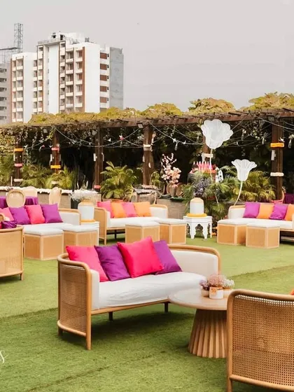 A vibrant outdoor setup for Devika and Harsh's celebration. The space is filled with colorful seating arrangements in shades of pink, purple, and orange, creating a cheerful and festive garden party atmosphere.
