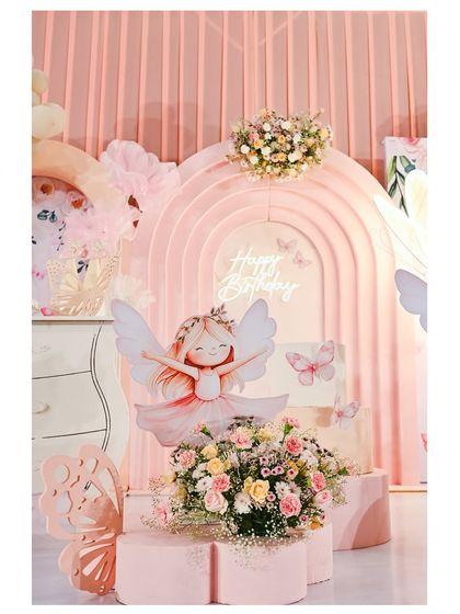 A beautiful fairy-themed dessert station. A large fairy cutout is the centerpiece, surrounded by fresh flowers and set against a multi-layered pink arch backdrop with a 'Happy Birthday' neon sign.