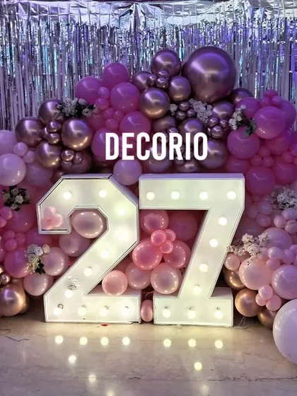 A fun and feminine 27th birthday decoration. This setup is built around large '27' marquee numbers, surrounded by a sea of pink and metallic balloons against a silver foil curtain backdrop.