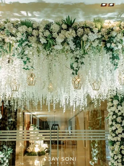 The floral entrance arch illuminated at night, with hanging lanterns casting a warm glow on the cascading white wisteria and dense floral pillars.