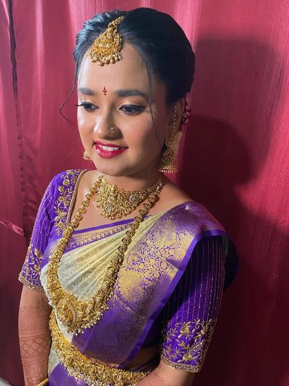A happy bride moments before her ceremony. The off-white and purple silk saree is neatly pleated, and the simple, elegant updo is finished with a maang tikka, creating a classic and radiant bridal look.