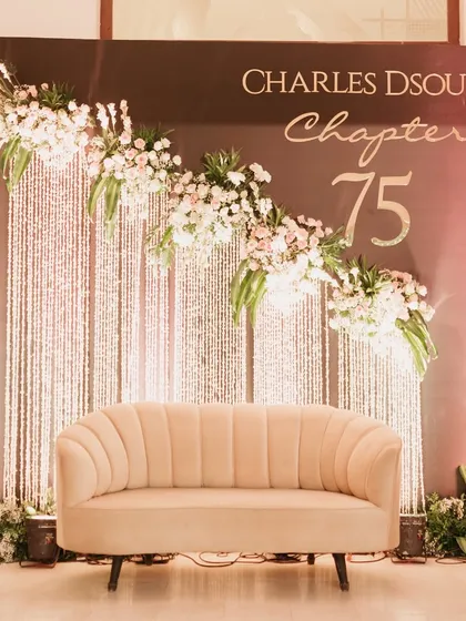 This elegant photo booth backdrop was designed for a 75th birthday party. It features a custom "Chapter 75" sign, a cascade of shimmering crystals, and beautiful floral arrangements, providing a perfect spot for memorable pictures.