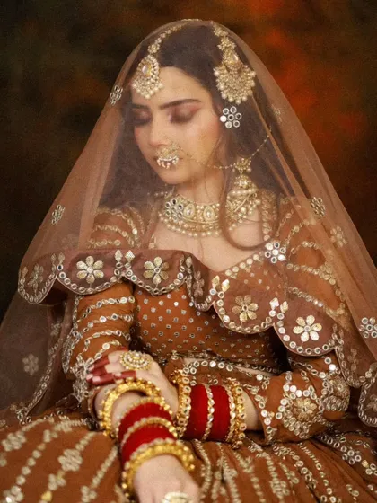 A bride in a beautiful earthy-toned lehenga with a sheer dupatta. The makeup is soft and natural, with a focus on warm, glowing skin.