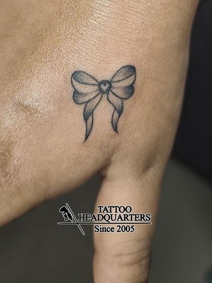 A small, shaded bow tattoo with a heart in the center, placed on the hand. A cute and delicate piece.
