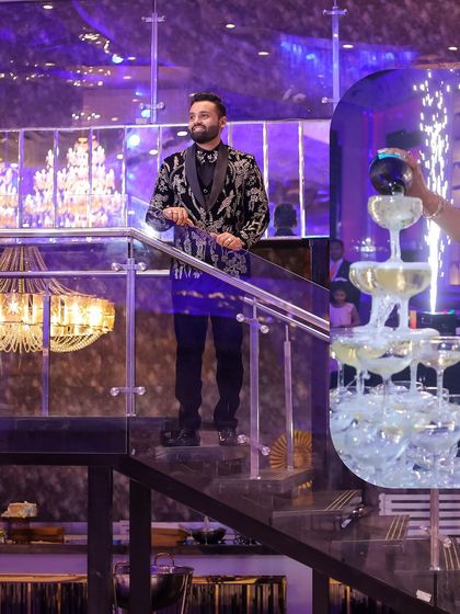 A stylish shot of the groom during the engagement party. He stands confidently on a staircase while a champagne tower is being poured, capturing the celebratory and luxurious feel of the event.