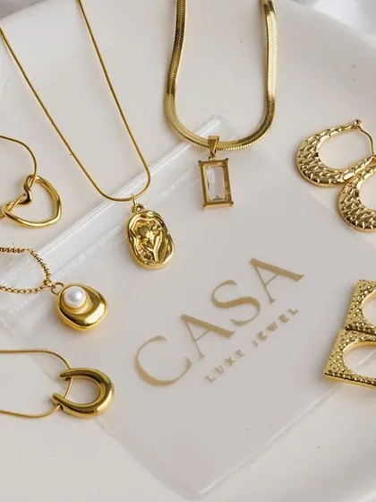 A collection of gold necklaces and earrings displayed on a branded plate against a soft white fabric. This group shot is great for showcasing a new collection or best-selling items.