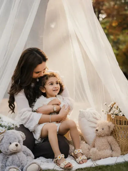 A mother kisses her daughter's head while they sit together in a sunlit teepee. A beautiful, warm portrait.