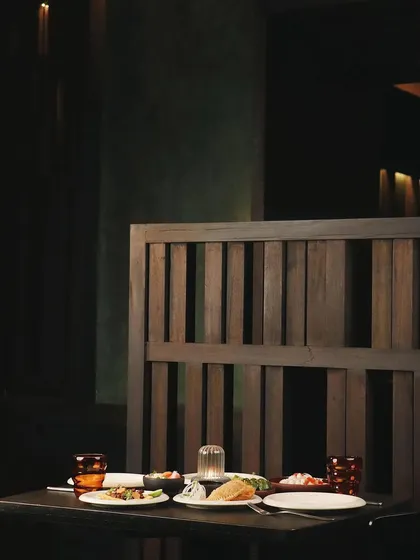 A table set for two at Otra. The dark wood of the furniture and partitions, combined with the soft, focused lighting, creates an intimate and sophisticated dining environment.