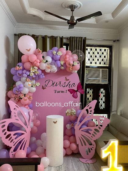 A whimsical butterfly-themed first birthday for 'Divisha'. The decor includes a pink arched backdrop, large butterfly wing cutouts, and a pink and purple balloon garland.
