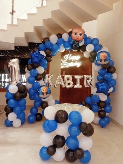 A Boss Baby themed first birthday with a blue, black, and white balloon arch, a "Happy Birthday" neon sign, and the child's name in foil balloons.