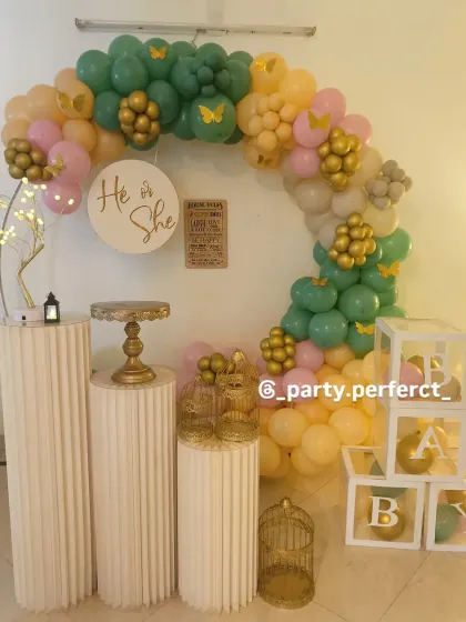 A stylish and modern gender reveal party setup. The "He or She" theme is captured with a circular backdrop and a beautiful balloon arch in sage green, peach, and gold, complemented by elegant white pedestals and 'B-A-B-Y' blocks.