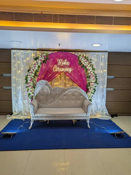 An elegant stage for a Roka ceremony at a banquet hall, designed with a floral ring, white and pink drapes, fairy lights, and a custom 'Roka Ceremony' neon sign.