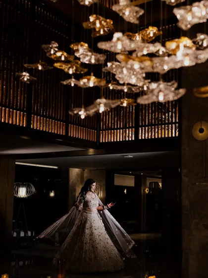 A dramatic portrait of the bride at her Sangeet, standing beneath a magnificent, modern chandelier. The grand architecture and lighting create a sense of scale and elegance.