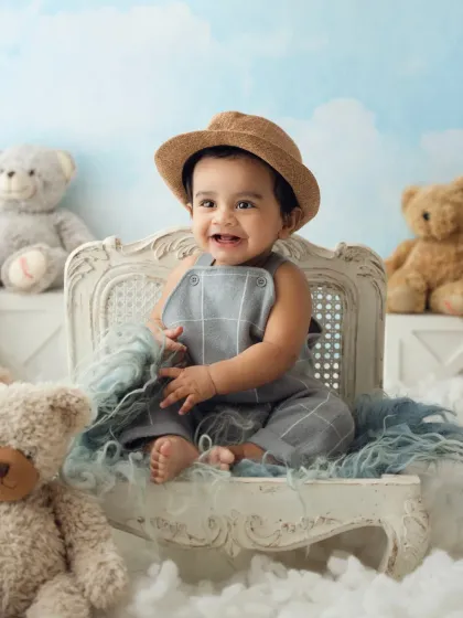 A smiling baby boy in a grey romper and hat, posing on a chair amidst a sea of clouds and teddy bears.