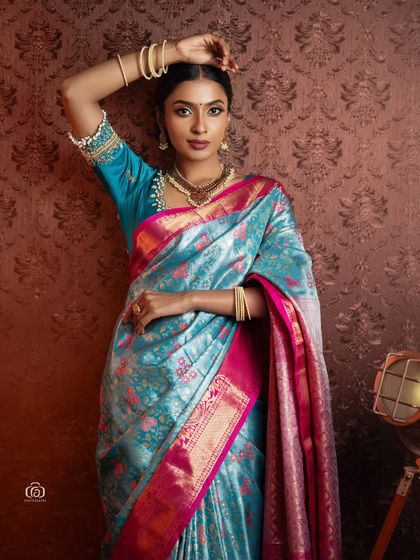 A different angle of the elegant blue and pink silk saree. This pose highlights the intricate embroidery on the sleeve and the graceful drape of the fabric, captured with warm studio lighting.