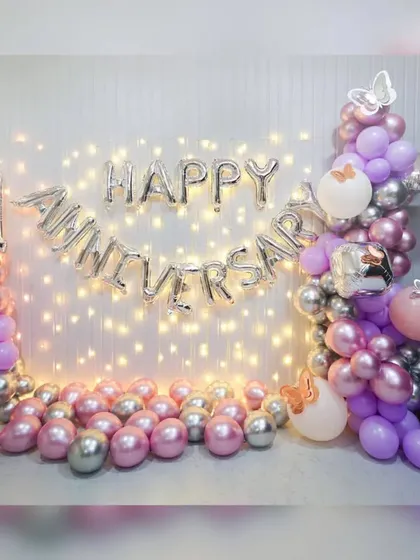 A first-anniversary decoration at home with a fairy light wall, 'Happy Anniversary' foil balloons, and a pink and silver balloon garland.
