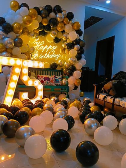 Celebrating a 75th birthday with a room full of joy. This home decoration features a balloon arch in black, gold, and white, large light-up marquee numbers, and dozens of balloons scattered on the floor for a festive feel.