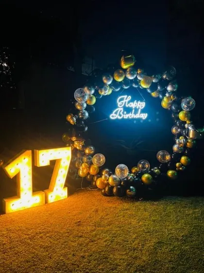 A dramatic nighttime birthday setup on a lawn, with a circular balloon frame, a neon sign, and large light-up marquee numbers.