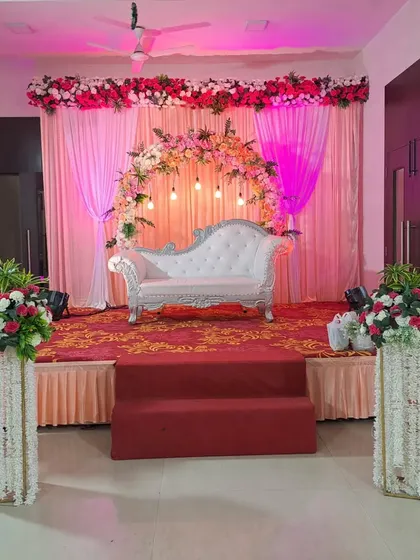 A complete stage setup inside a hall, with a floral arch, a white chaise lounge, and matching floral pillars, ideal for an engagement or roka ceremony.