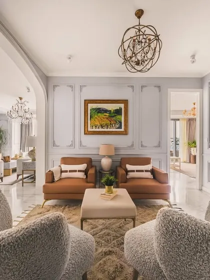 This is a perspective of a neoclassical living room, showcasing a masterful blend of classic elegance and modern comfort. The plush, textured armchairs in the foreground invite you into a space defined by symmetrical design, ornate wall moldings, and a grand, gilded mirror that amplifies the room's luxurious feel.