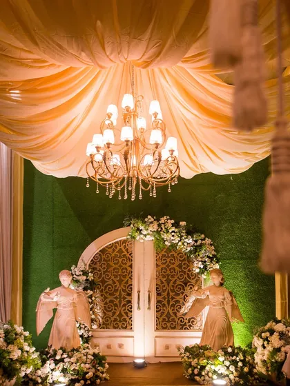 A dramatic entrance for a cocktail event, with a grand doorway framed by angel statues, white florals, and a draped ceiling with a chandelier.