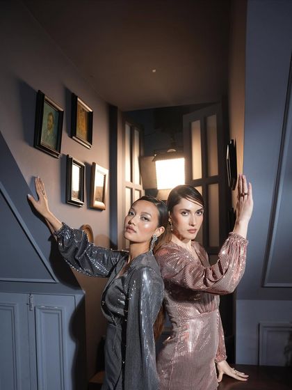 Two models styled in coordinating metallic dresses, one silver and one rose gold. The back-to-back pose in a dimly lit hallway creates a sense of mystery and glamour.