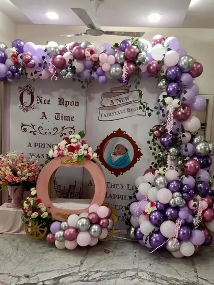 This "Once Upon a Time" setup was designed for a baby girl's welcome home party. It features a custom fairytale book backdrop, a princess carriage prop for photos, and a lush balloon garland in shades of purple, pink, and silver.
