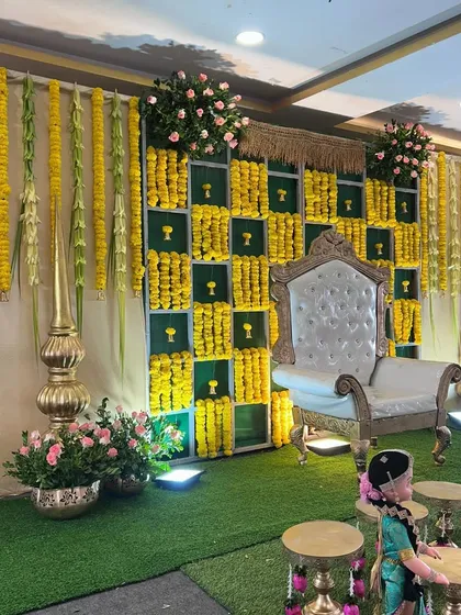 A beautiful and traditional baby shower stage decoration. The setup features a stunning backdrop of green panels and yellow marigold flowers, an ornate white throne, and classic Indian decorative elements.
