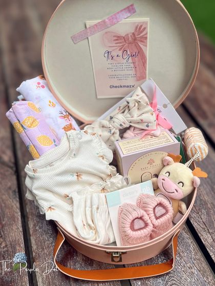 Opening the hat box reveals a curated collection of essentials for a baby girl, including a floral onesie, lemon-print swaddle, soft booties, and a plush giraffe toy.