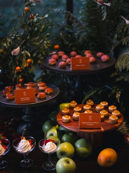 A variety of miniature desserts, including vanilla choux buns and fruit tarts, are displayed on multi-level stands. The small labels add a professional and informative touch.