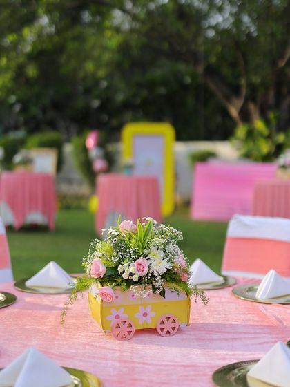 A charming floral centerpiece in a custom yellow wagon-shaped vase. This detail from the "Sunshine & Pastel Rainbows" party adds a whimsical and personal touch to the table decor.