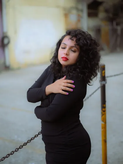 A candid-style portrait with a soft, out-of-focus urban background. The focus is on the model's expression and the texture of her curly hair.