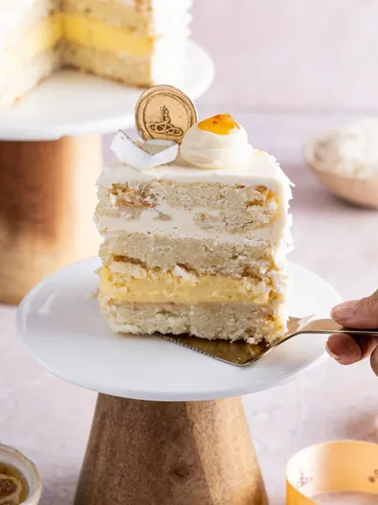A slice of our tropical Coconut and Passionfruit cake on a dessert stand. This cake features layers of fluffy coconut sponge, a bright passion fruit cremeux, and a smooth coconut buttercream, perfect for a taste of summer.