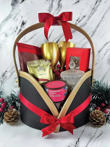 A holiday hamper designed for celebration. This basket features golden goblets, cocktail mix, and cranberry treats, all tied together with a festive red ribbon against a classic marble background.