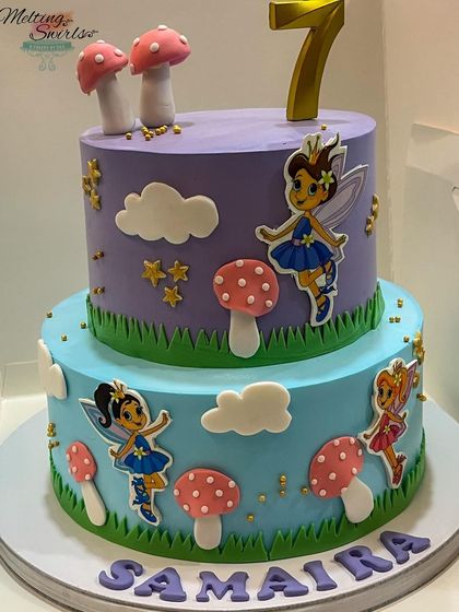 A whimsical two-tiered fairy cake decorated with edible fairy prints and handmade fondant mushrooms. The purple and blue colors create an enchanted forest feel.