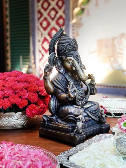 An antique-finish Ganesha idol placed among ceremonial offerings. The setup includes silver trays with fresh flower petals and vibrant red carnations, invoking blessings for the new beginning.